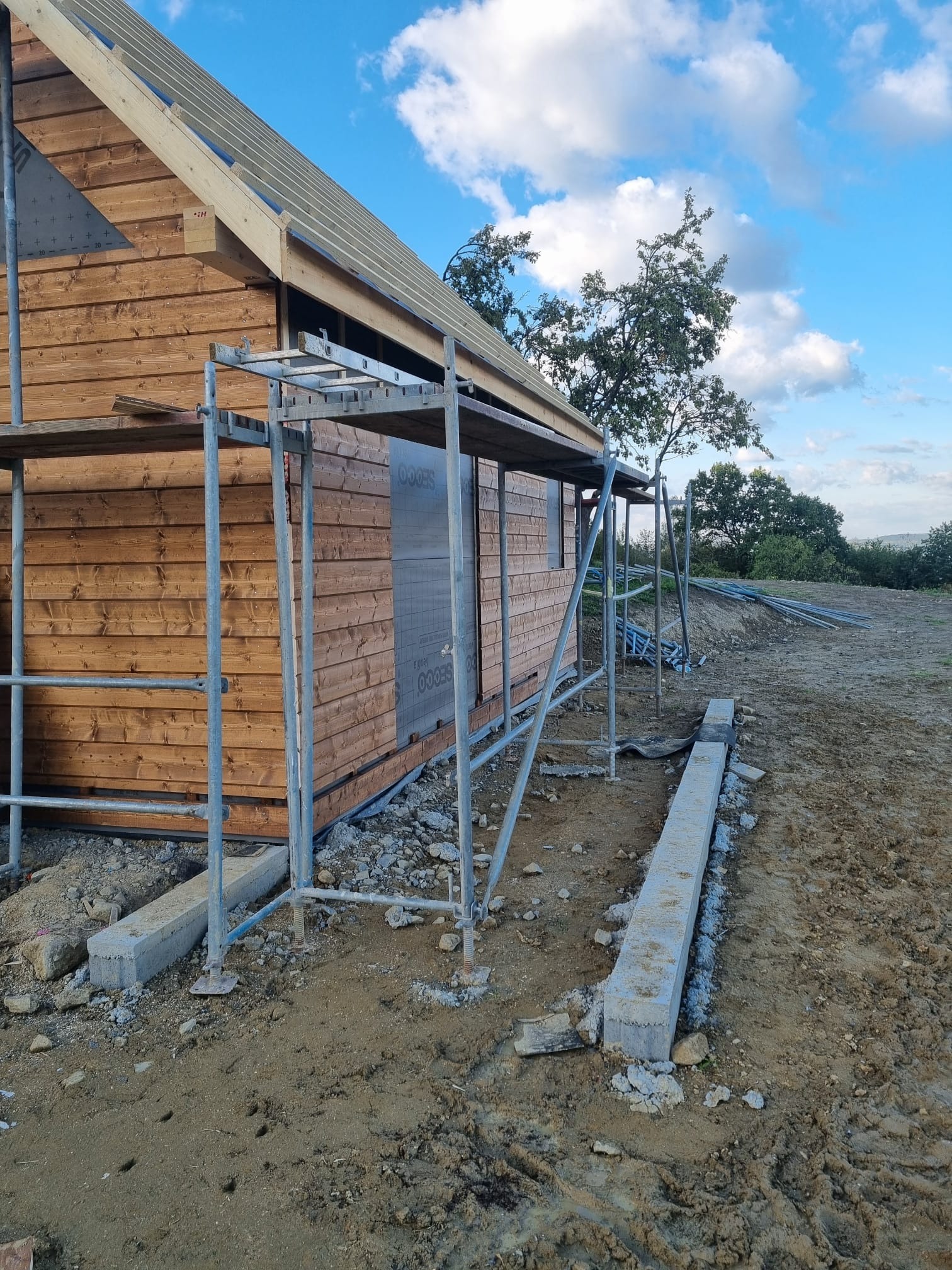 Surowy stan domu szkieletowego z drewnianą elewacją, widoczne rusztowania i przygotowane fundamenty na tle błękitnego nieba z chmurami.