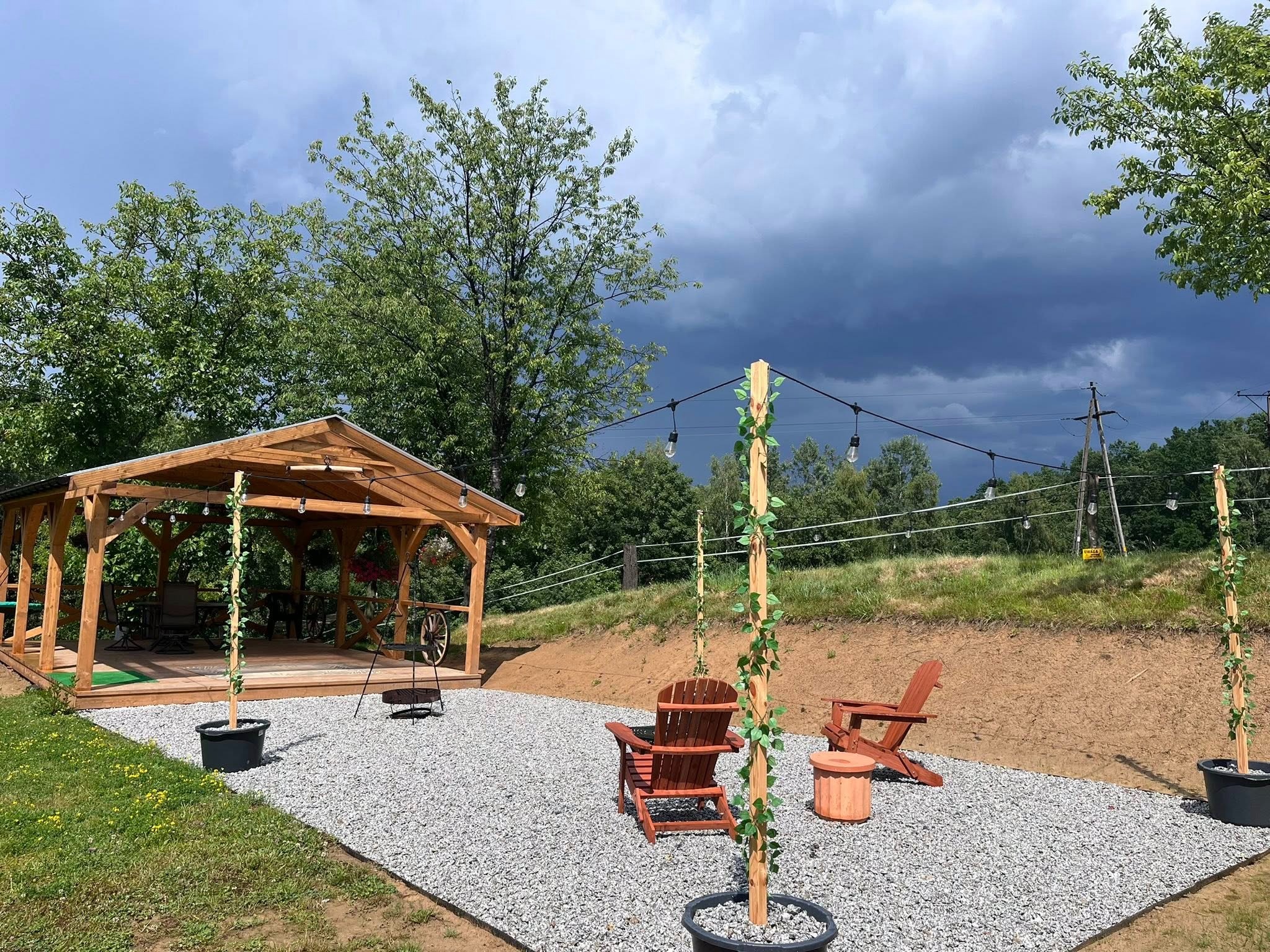 Drewniany taras z zadaszeniem, krzesła Adirondack i dekoracyjne słupki z bluszczem na tle burzowego nieba. Elementy drewniane ocieplone światłem, kamienny żwir.