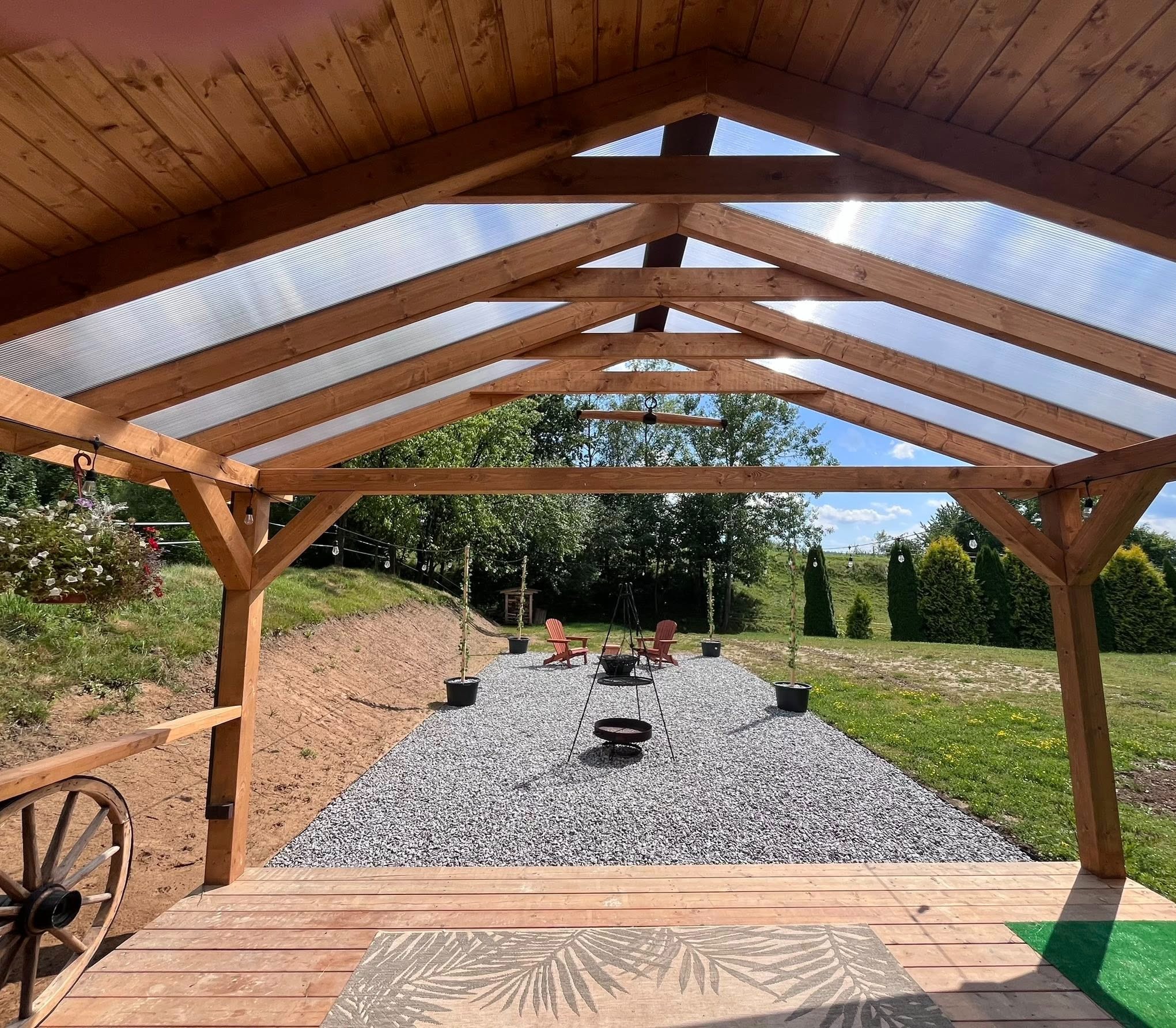 Zadaszona, drewniana altana z transparentnym dachem. Widok na ogród z miejscem na ognisko i relaks. Na pierwszym planie drewniany podest i dekoracyjny dywan.