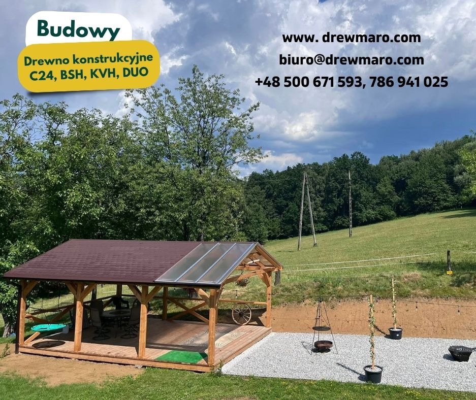 Drewniana altana z brązowym dachem i przezroczystym panelem, otoczona zielenią. Widoczne elementy dekoracyjne, takie jak koło od wozu i wiszące donice.