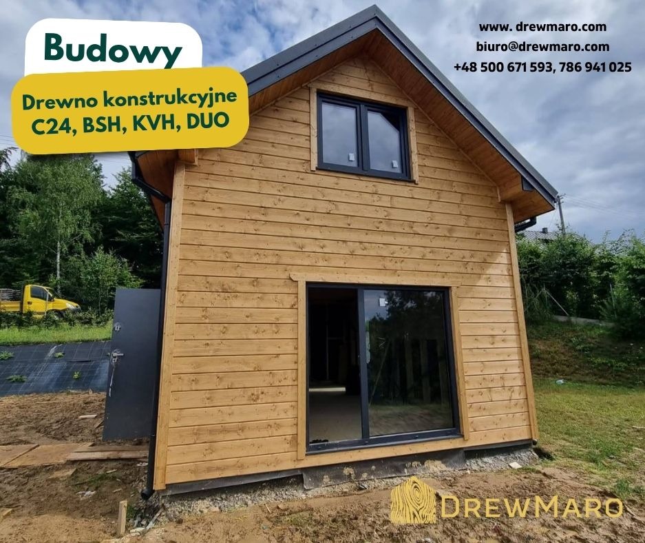 Gotowy domek szkieletowy z jasnego drewna, z czarną stolarką okienną i drzwiami. Widoczny fragment działki i auto dostawcze w tle. Budowa w toku.