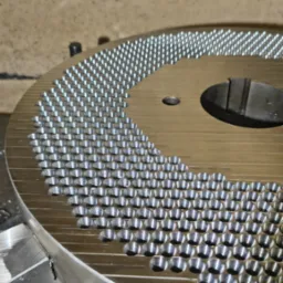 Metalowy dysk z rzędami precyzyjnie wywierconych otworów, widoczne ślady obróbki, surowy detal po procesie wiercenia, jasne oświetlenie.