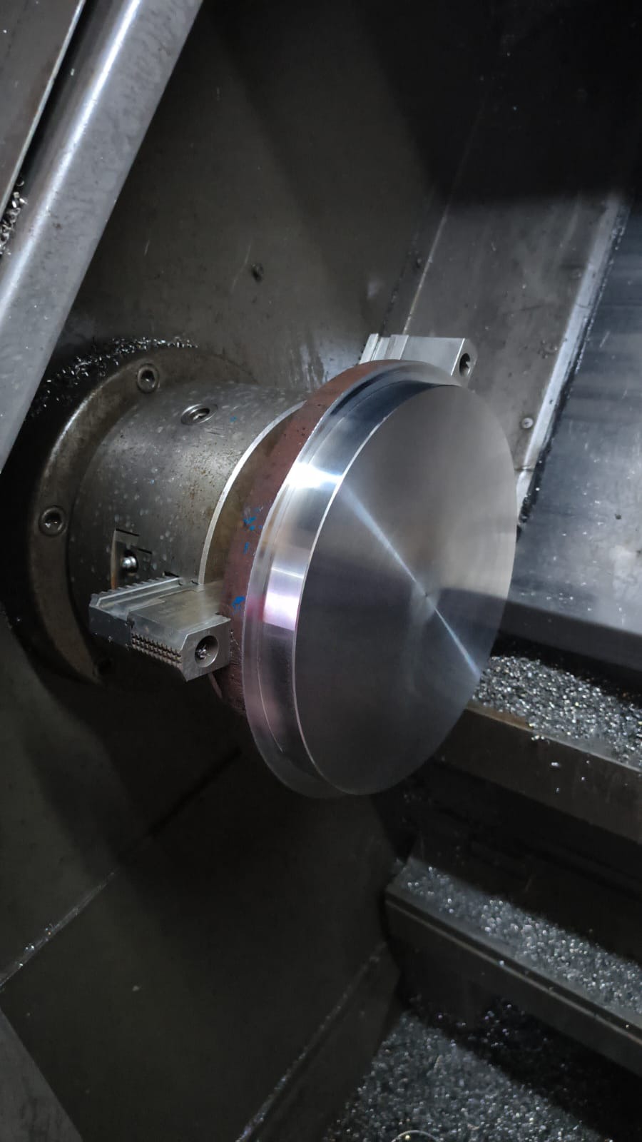Metalowy dysk w uchwycie tokarki, z widocznymi wiórami, co sugeruje proces obróbki skrawaniem. Oświetlenie podkreśla gładką powierzchnię detalu.