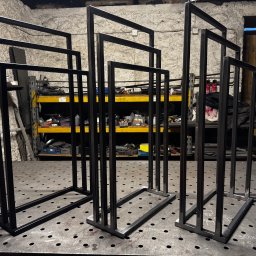 Loft Weld - Surowe, czarne, metalowe ramy o prostokątnym przekroju, ustawione w warsztacie na perforowanym stole spawalniczym. W tle półki z narzędziami i materiałami.