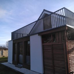 Loft Weld - Nowoczesny dom z balkonem zabezpieczonym czarną, metalową balustradą. Widoczne drewniane elementy konstrukcyjne i jasna elewacja. Ujęcie z dołu, w tle błękitne niebo.