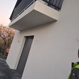 Loft Weld - Nowoczesny balkon z czarną, metalową balustradą, przylegający do białego budynku z szarą roletą. Widok z dołu, podkreślający minimalistyczny design i geometryczne kształty.