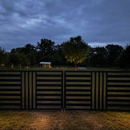 Loft Weld - Nowoczesna, dwuskrzydłowa brama z ciemnych, poziomych profili z pionowymi akcentami, osadzona w betonowych słupach z ciepłym oświetleniem. W tle oświetlone drzewo i budynek o zmierzchu.