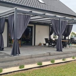 Loft Weld - Nowoczesny dom z dużym, zadaszonym tarasem, wyposażonym w ciemnoszare zasłony i przezroczysty dach. Na patio stół z krzesłami, obok trawnik z rabatą lawendy. W tle pagórki.