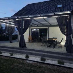 Loft Weld - Zadaszenie tarasu z ciemnego metalu i przezroczystego poliwęglanu, z podświetleniem LED i szarymi zasłonami, na brukowanym patio z meblami ogrodowymi, o zmierzchu.