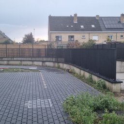 Mballu - Ciemnoszara kostka brukowa na podjeździe, otoczona niskim murem i czarnym metalowym ogrodzeniem. W tle domy jednorodzinne pod pochmurnym niebem.