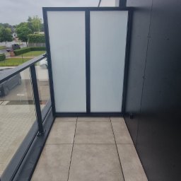 Mballu - Nowoczesny balkon z szklaną balustradą i panelami, wykończony jasnymi płytkami. Widok na zewnątrz, szara kolorystyka i minimalistyczny design.