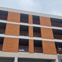 Mballu - Nowoczesny budynek z balkonami zabezpieczonymi metalowymi balustradami w kolorze miedzianym. Osoba na jednym z balkonów. Elewacja z cegły w dwóch odcieniach.