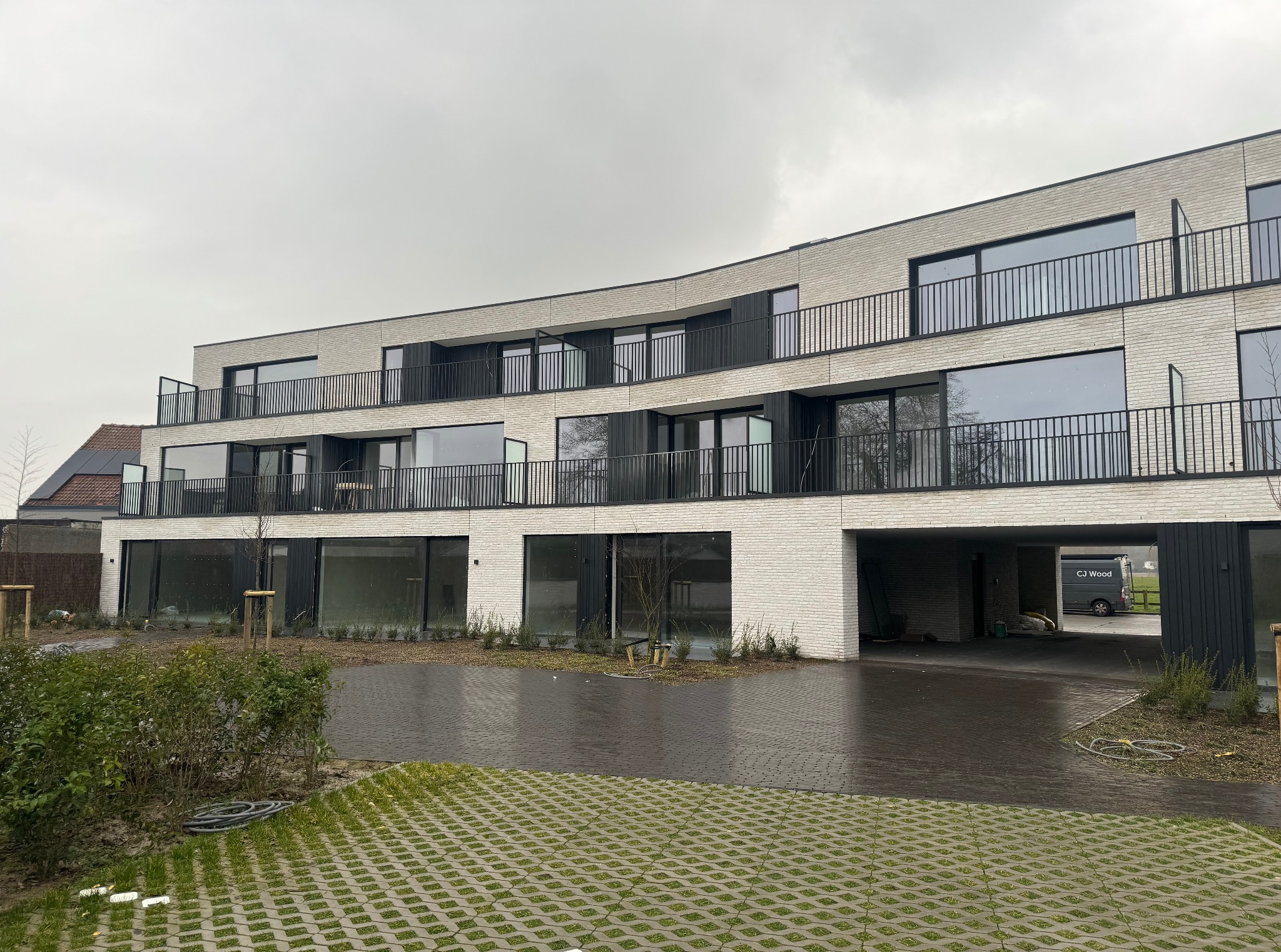 Nowoczesny budynek wielorodzinny z balkonami zabezpieczonymi czarnymi, metalowymi balustradami. Widoczna kostka brukowa na podjeździe i zieleń wokół.