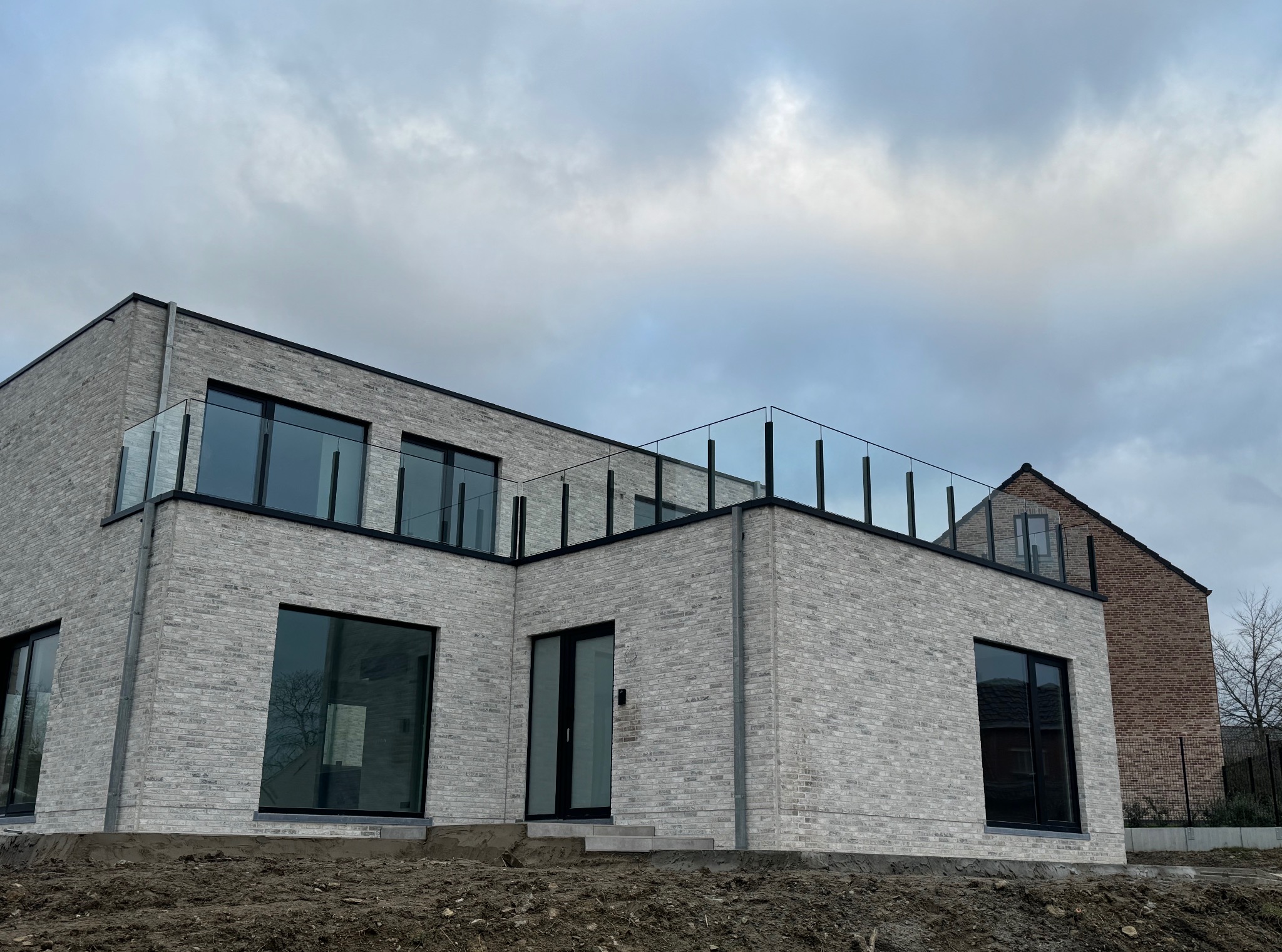 Nowoczesny, dwukondygnacyjny dom z jasnej cegły z szklanymi balustradami na płaskim dachu, widok z dołu na tle pochmurnego nieba.