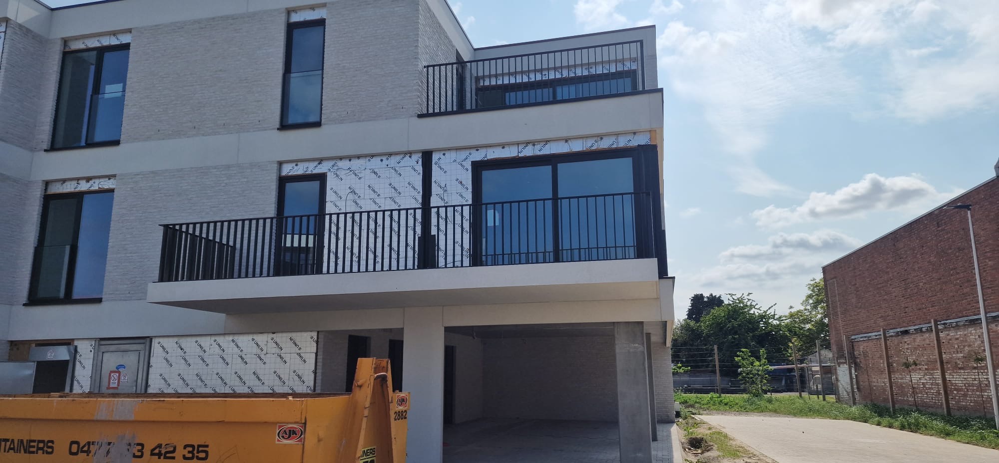 Nowoczesny budynek z balkonami zabezpieczonymi czarnymi, metalowymi balustradami. Widoczny kontener budowlany u dołu kadru.