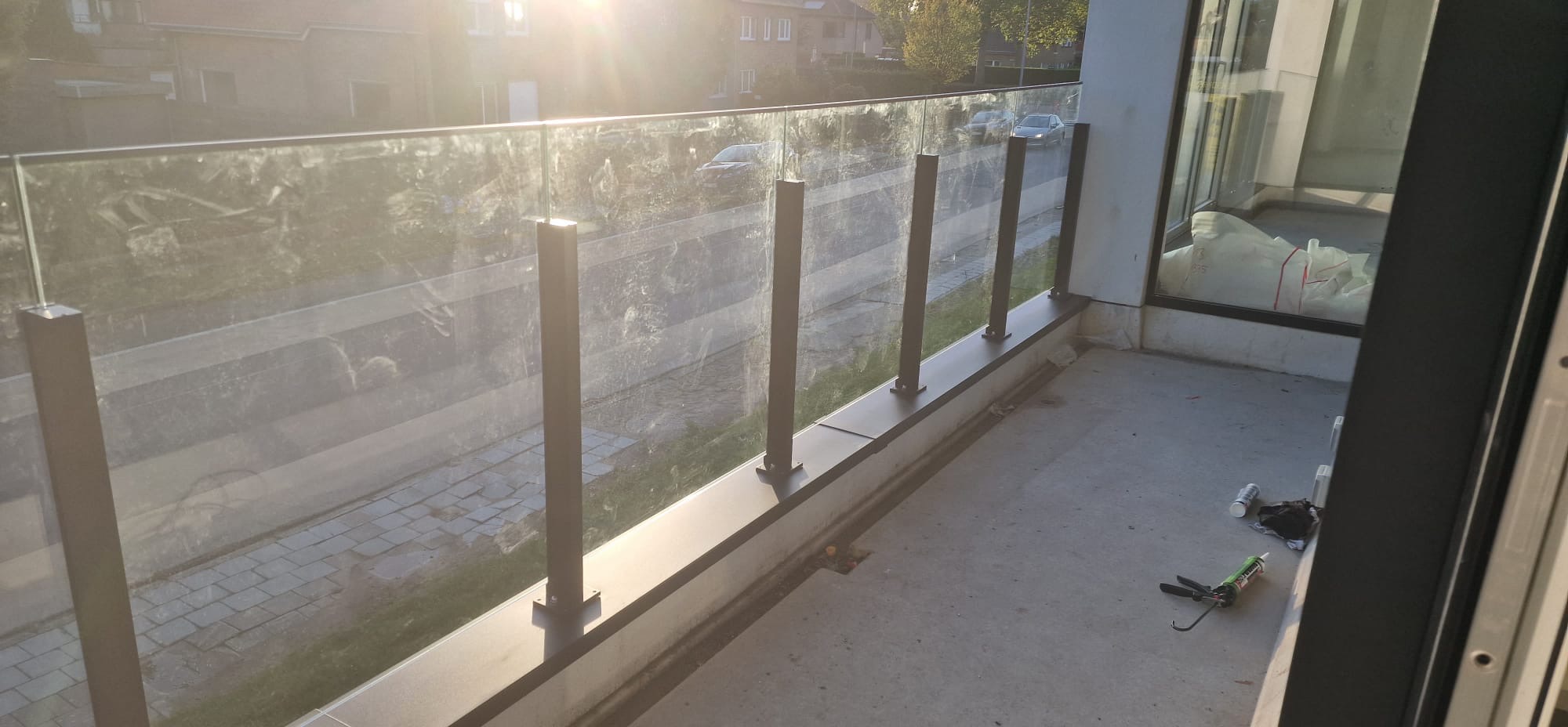 Balkon z nowoczesną szklaną balustradą z ciemnymi, metalowymi słupkami, widok na ulicę. Narzędzia montażowe na posadzce sugerują świeży montaż.