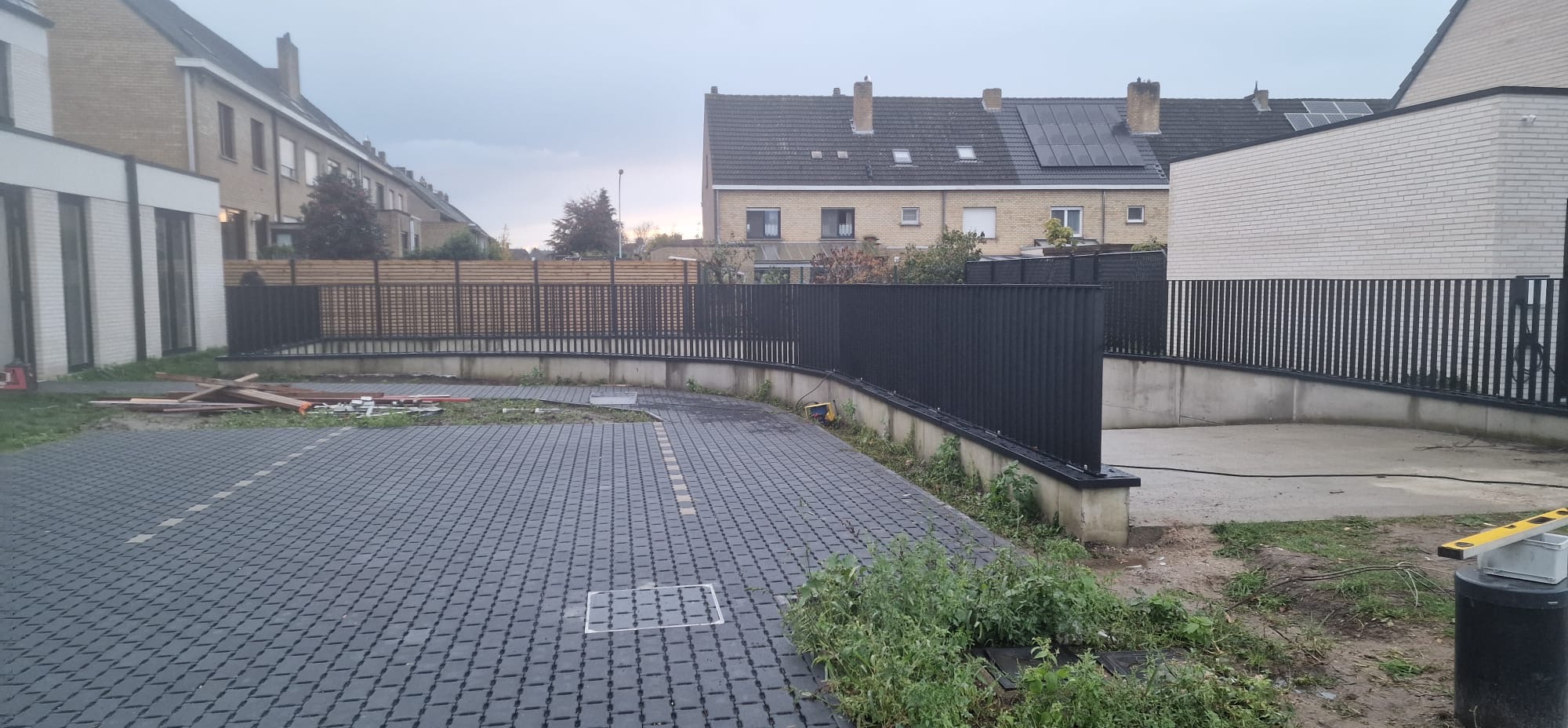 Ciemnoszara kostka brukowa na podjeździe, otoczona niskim murem i czarnym metalowym ogrodzeniem. W tle domy jednorodzinne pod pochmurnym niebem.