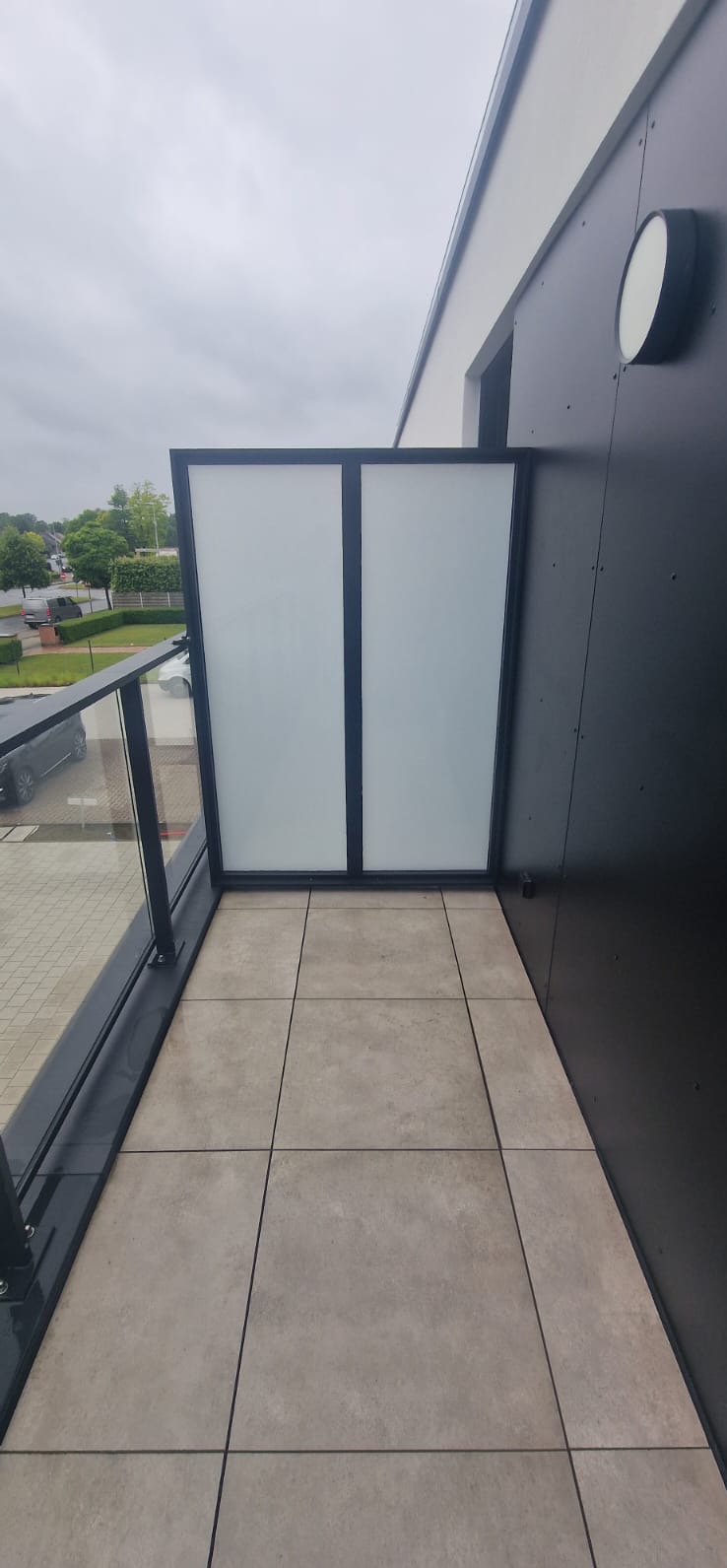 Nowoczesny balkon z szklaną balustradą i panelami, wykończony jasnymi płytkami. Widok na zewnątrz, szara kolorystyka i minimalistyczny design.