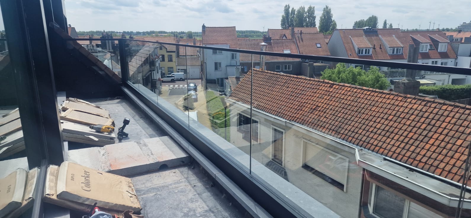 Szklana balustrada z czarną ramą na dachu budynku, widok na miasto. Częściowo widoczne narzędzia i materiały montażowe. Słoneczny dzień.