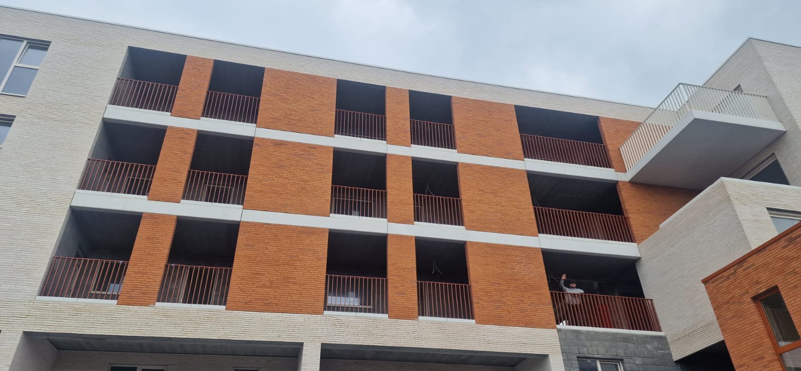 Nowoczesny budynek z balkonami zabezpieczonymi metalowymi balustradami w kolorze miedzianym. Osoba na jednym z balkonów. Elewacja z cegły w dwóch odcieniach.