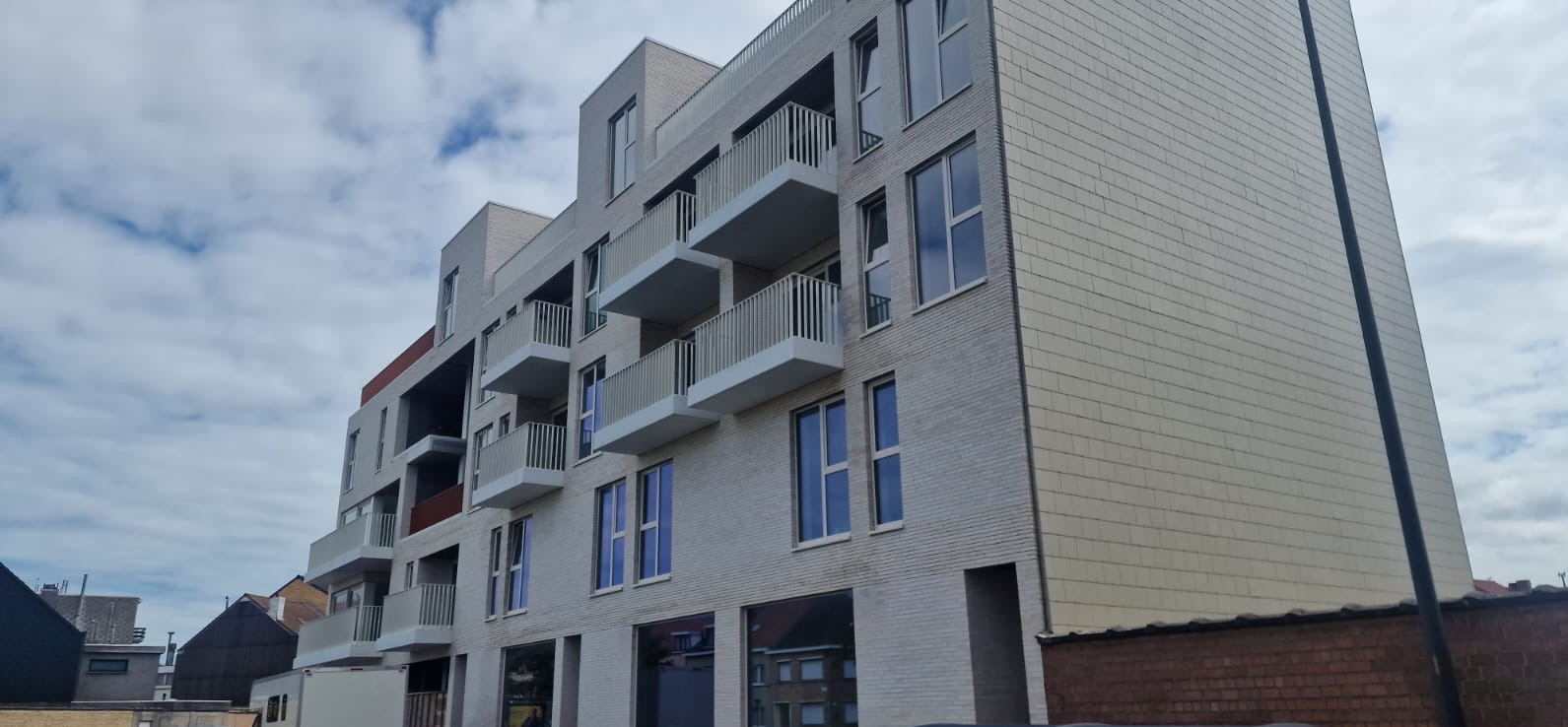 Elewacja budynku wielorodzinnego z balkonami o białych, metalowych barierkach. Widok z dołu ukazuje okna i ceglaną fasadę w odcieniach szarości.