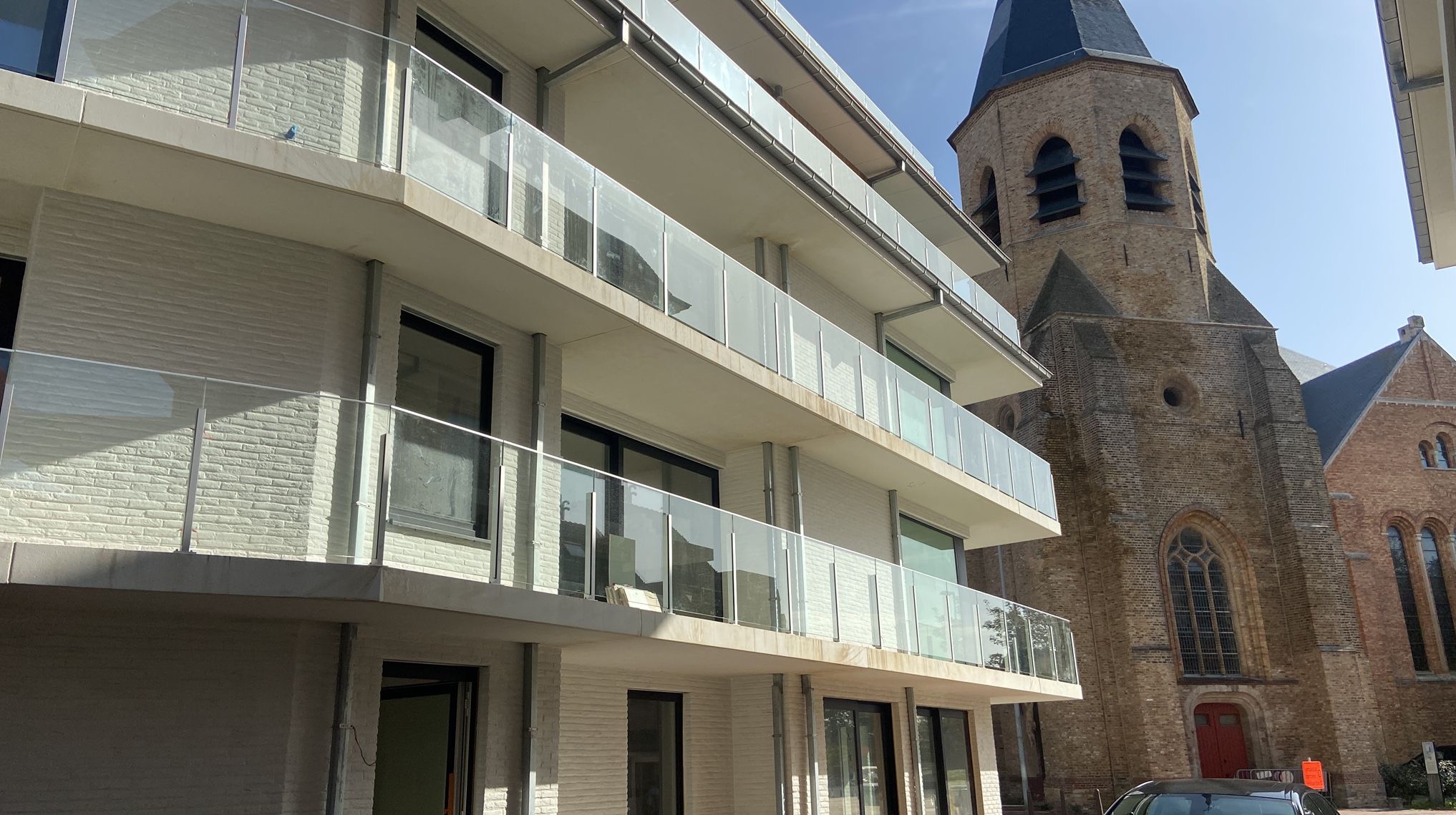 Nowoczesny budynek z białej cegły z balkonami zabezpieczonymi szklanymi balustradami na tle starego kościoła z cegły. Jasne, słoneczne ujęcie.