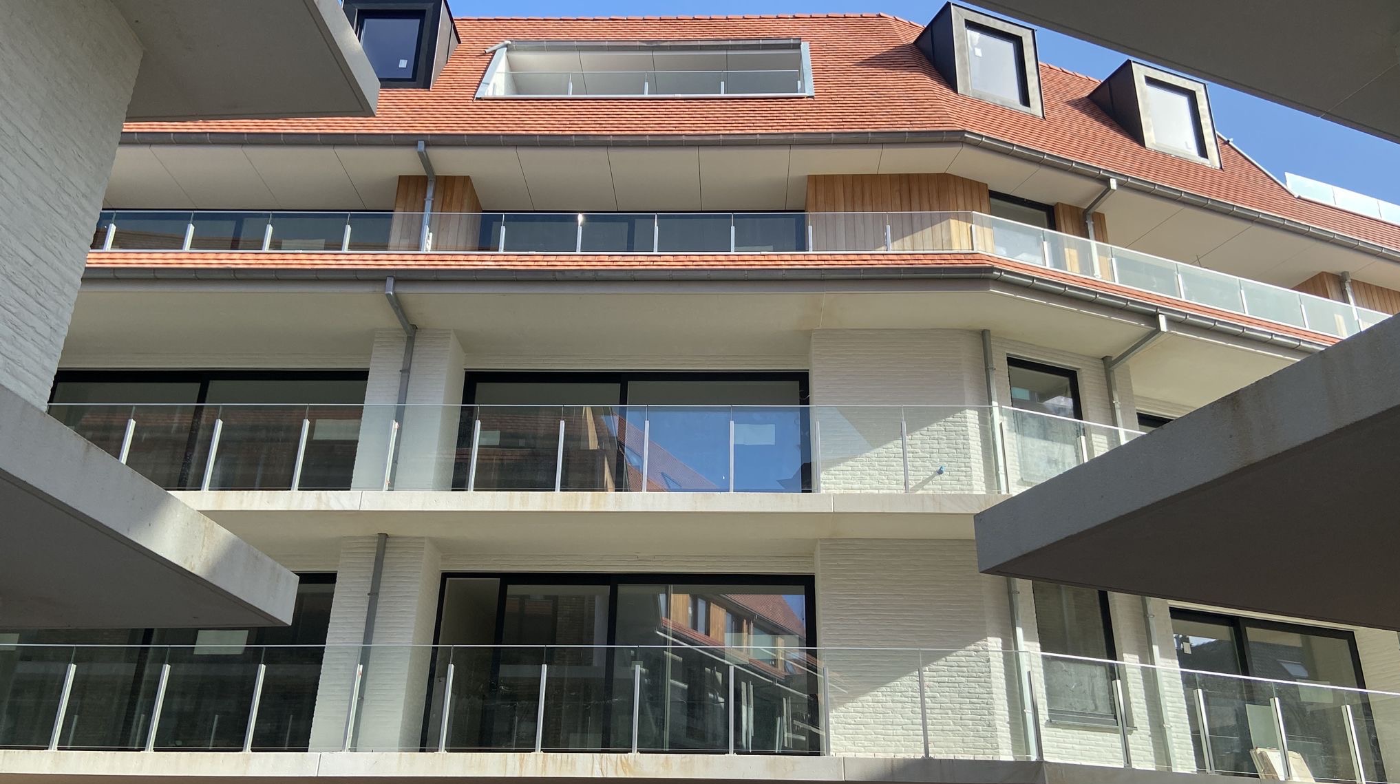 Elewacja budynku z balkonami i szklanymi balustradami. Ujęcie z dołu, widoczny fragment dachu z oknami. Jasna cegła, nowoczesny design.