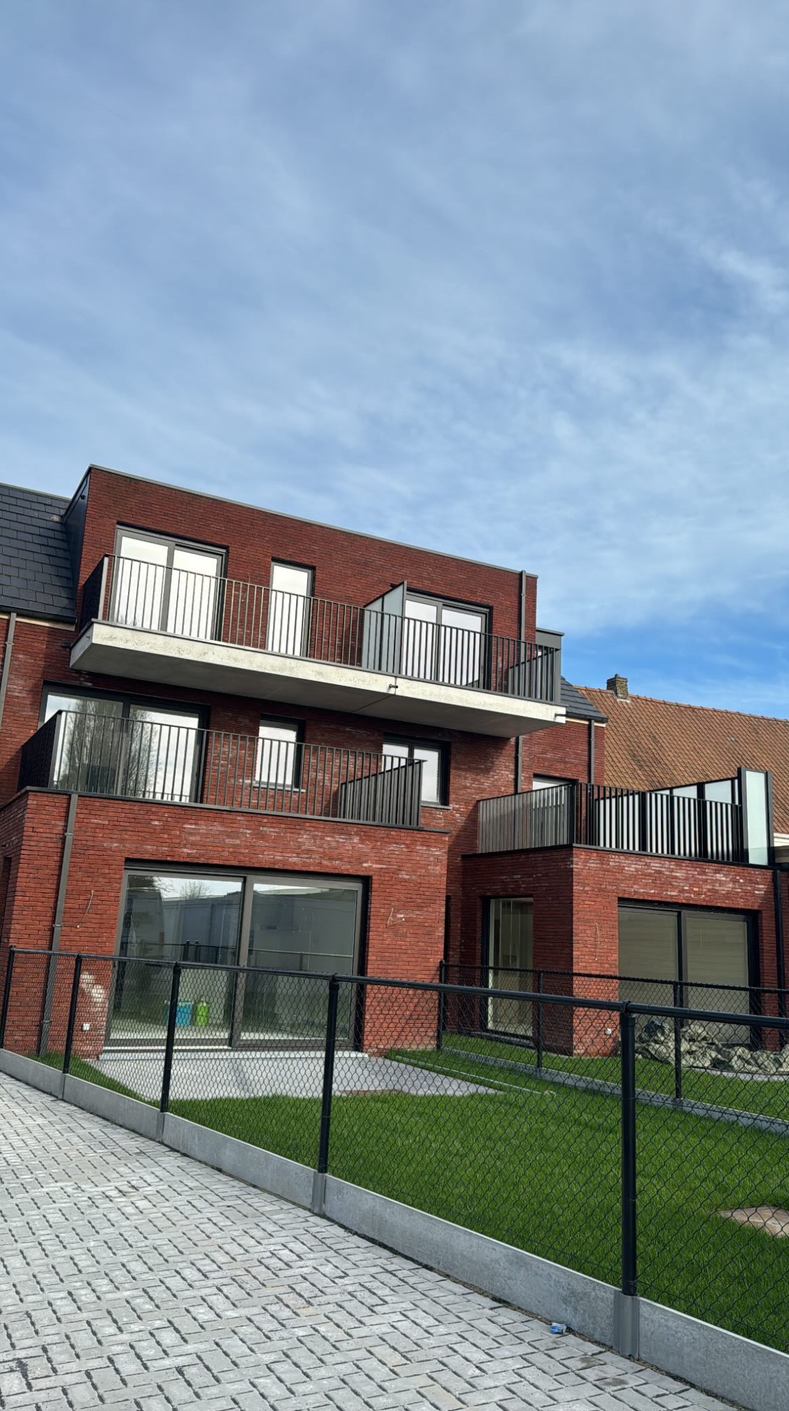 Nowoczesny budynek z cegły z balkonami z metalowymi balustradami. Kostka brukowa prowadzi do ogrodzenia z siatki. Błękitne niebo w tle.