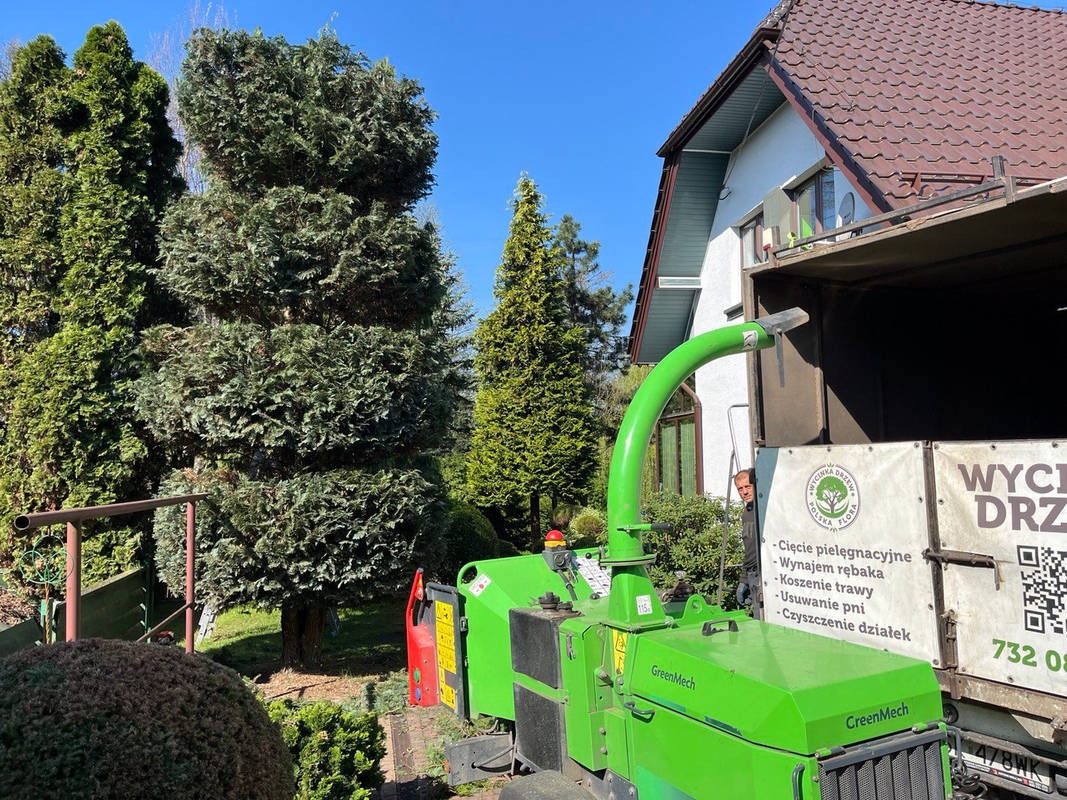 Rębarka GreenMech przy ciężarówce z napisem 'Wycinka Drzew', w tle drzewa iglaste i dom. Widoczny pracownik. Usługa wycinki drzew w ogrodzie.