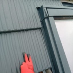 nowydach - Montaż okna dachowego z szarą blachodachówką, widoczne rękawice robocze i wkrętarka. Detal obróbki blacharskiej przy oknie.
