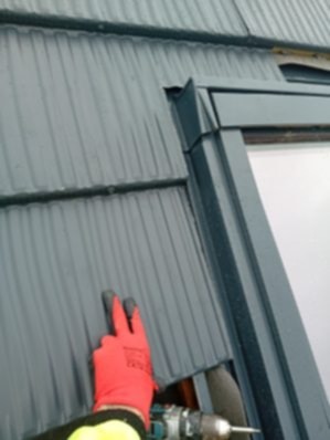 Montaż okna dachowego z szarą blachodachówką, widoczne rękawice robocze i wkrętarka. Detal obróbki blacharskiej przy oknie.