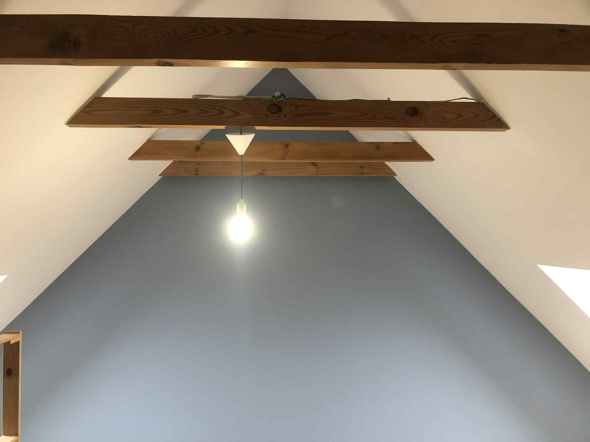 Poddasze z drewnianymi belkami stropowymi, pomalowane ściany na biało i szaro-niebiesko, wisząca żarówka. Widok z dołu.