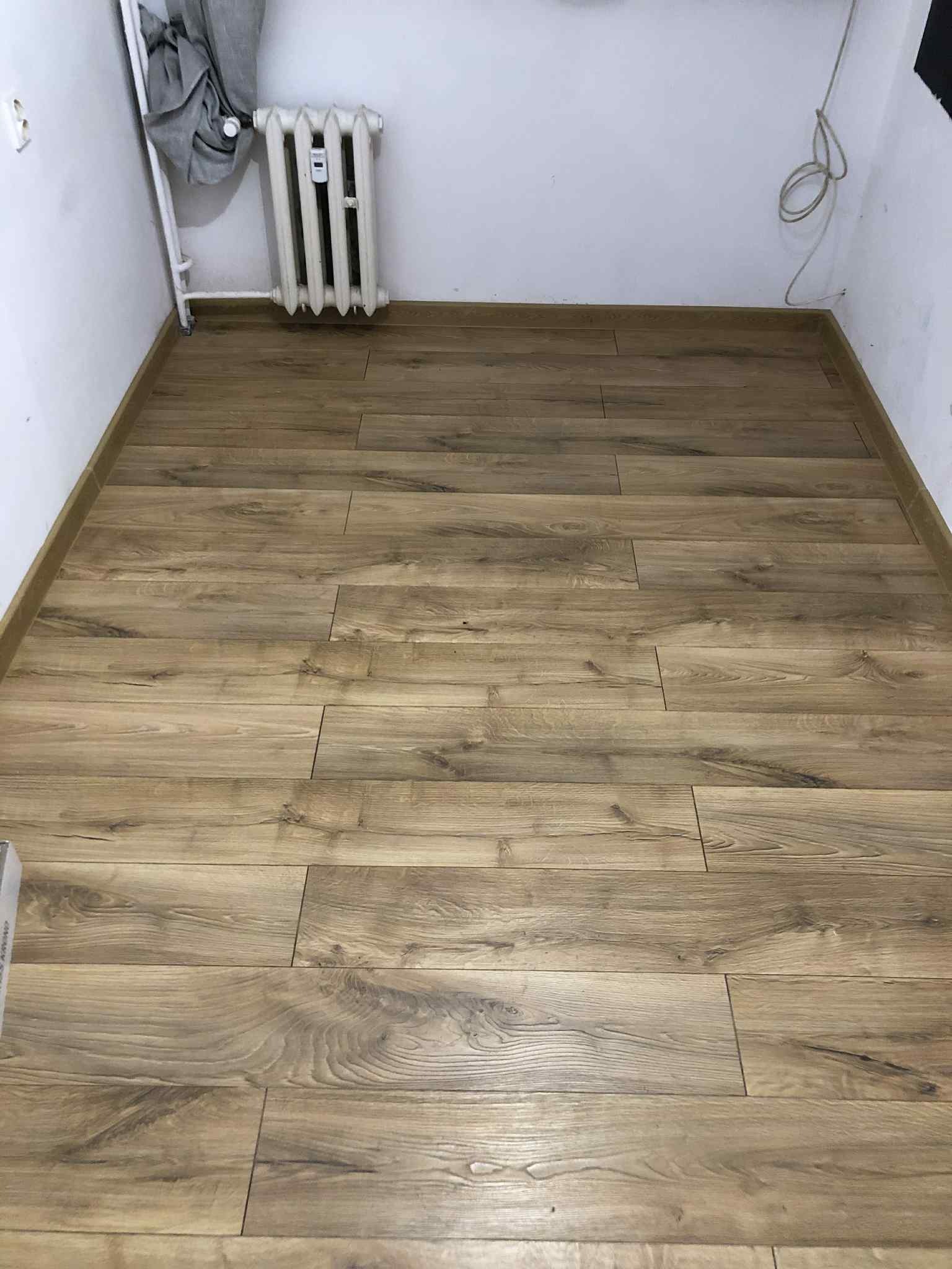 Ułożone panele podłogowe w pokoju z grzejnikiem i listwami przypodłogowymi. Widoczna faktura drewna i jasne oświetlenie.
