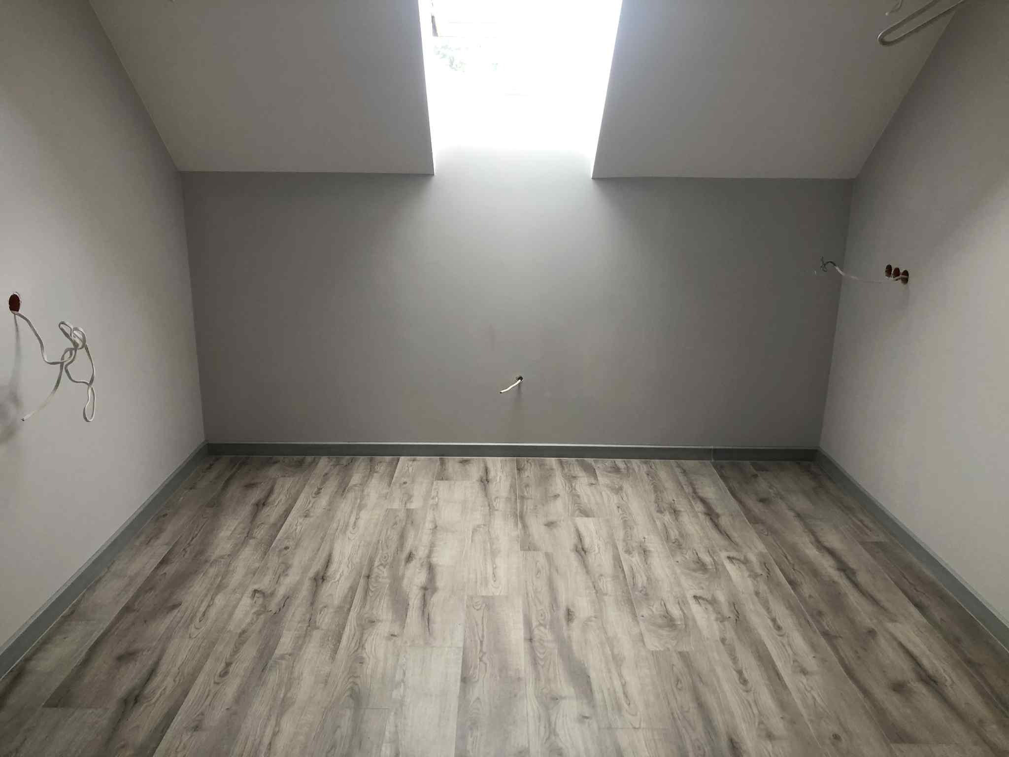 Poddasze z ułożonymi panelami podłogowymi w odcieniach szarości, widoczne okna dachowe i fragment instalacji elektrycznej na ścianie.