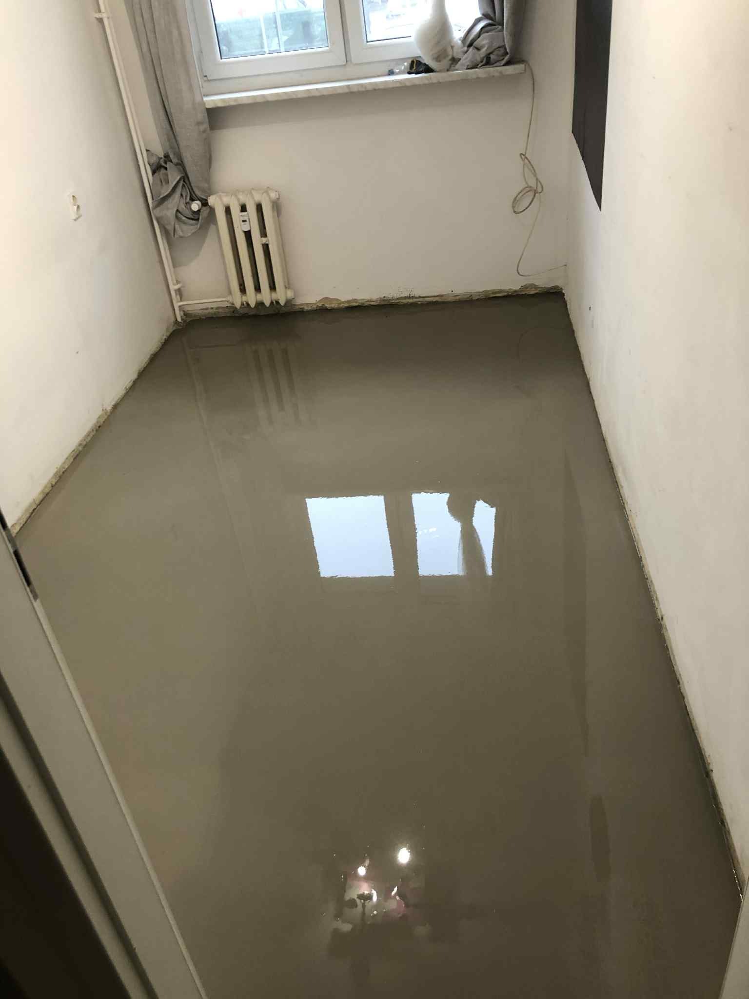 Wylana, mokra wylewka samopoziomująca na podłodze w pokoju z grzejnikiem i oknem. Widoczne odbicie okna w mokrej powierzchni. Szare ściany.