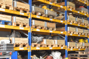 Magazyn z częściami do hydrauliki siłowej: regały wypełnione paletami i podzespołami do maszyn budowlanych i przemysłowych. Żółto-niebieska konstrukcja regałów.