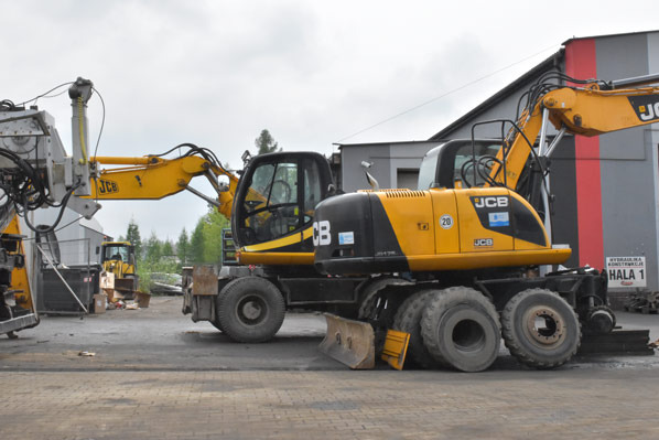 Żółte koparki JCB na placu przed budynkiem z szyldem, w tle widoczny inny sprzęt budowlany i zieleń.