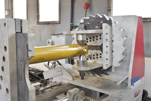 Żółty siłownik hydrauliczny w maszynie serwisowej z ostrzami tnącymi, gotowy do naprawy lub regeneracji. Widoczne narzędzia i elementy konstrukcyjne.