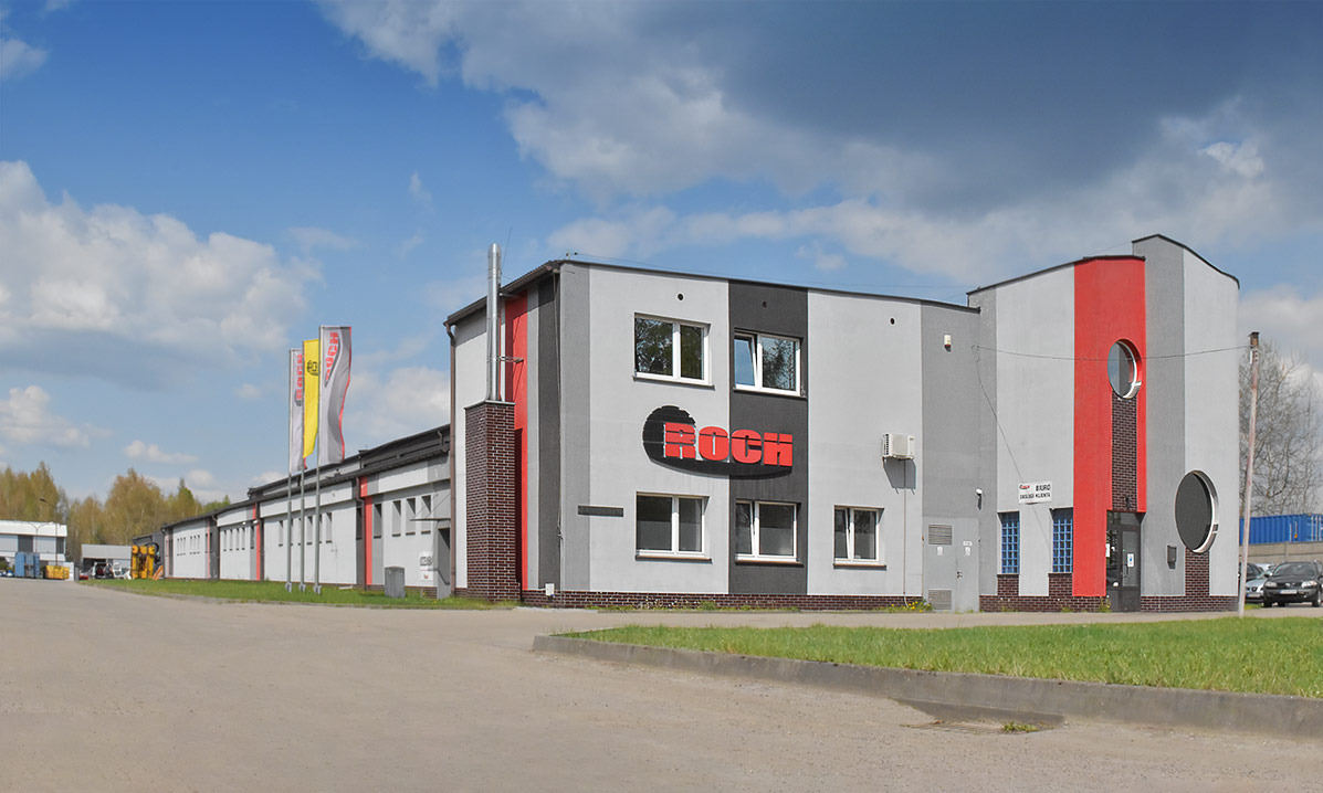 Budynek zakładu serwisowego maszyn z logo firmy Roch, otoczony zielenią i placem manewrowym, w słoneczny dzień. Widoczne flagi firmowe i zaparkowane samochody.