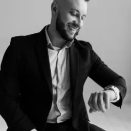 Czarno-białe zdjęcie uśmiechniętego mężczyzny w garniturze, spoglądającego na zegarek na ręce. Elegancki wygląd, biznesowy styl.