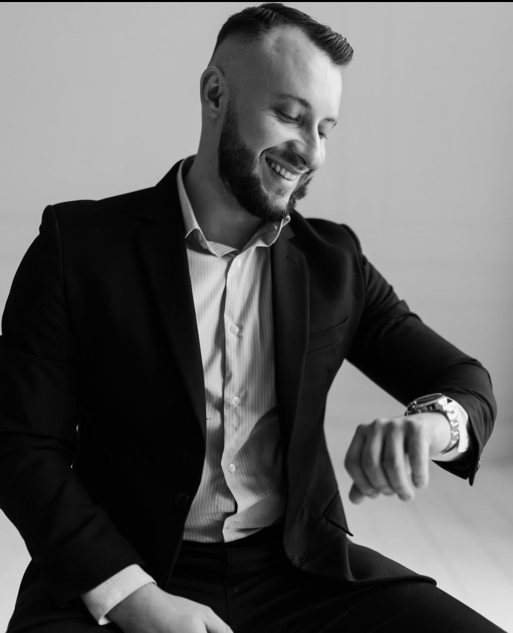 Czarno-białe zdjęcie uśmiechniętego mężczyzny w garniturze, spoglądającego na zegarek na ręce. Elegancki wygląd, biznesowy styl.