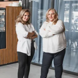 Dwie kobiety w białych koszulach stojące na tle okna i menu. Kobieta po lewej ma dżinsy i mokasyny, a kobieta po prawej ma spodnie i buty.