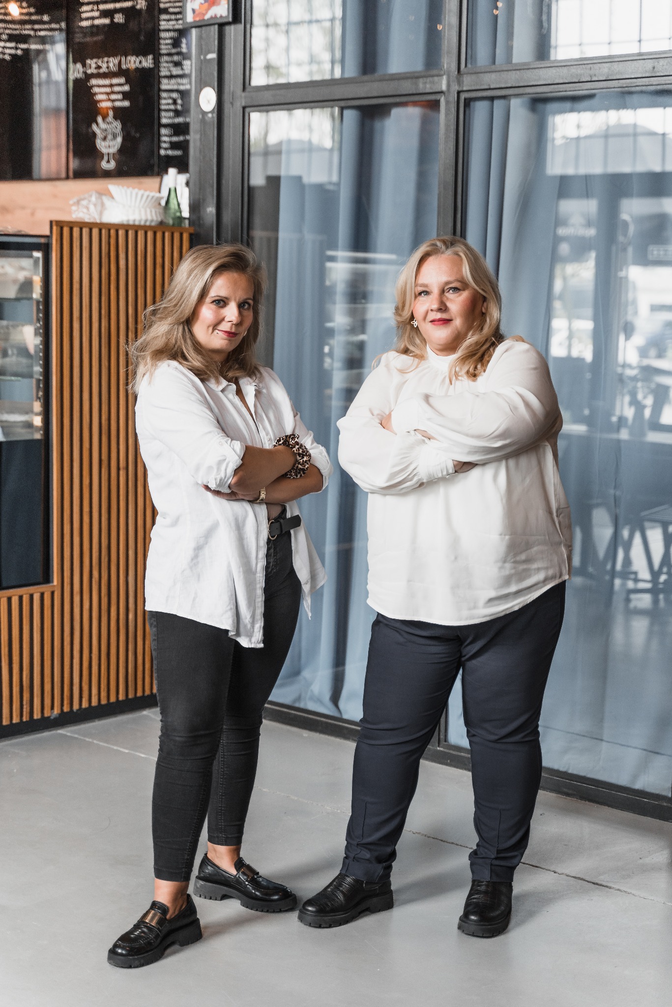 Dwie kobiety w białych koszulach stojące na tle okna i menu. Kobieta po lewej ma dżinsy i mokasyny, a kobieta po prawej ma spodnie i buty.