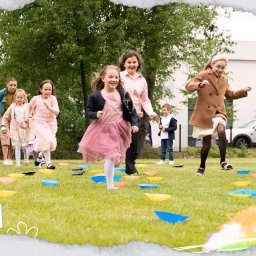 BV3 Video - Grupa dzieci biegnie po trawie po kolorowych podkładkach. Widoczny kamerzysta nagrywający wydarzenie. Ujęcie z niskiego kąta, plenerowe, z zielenią w tle.