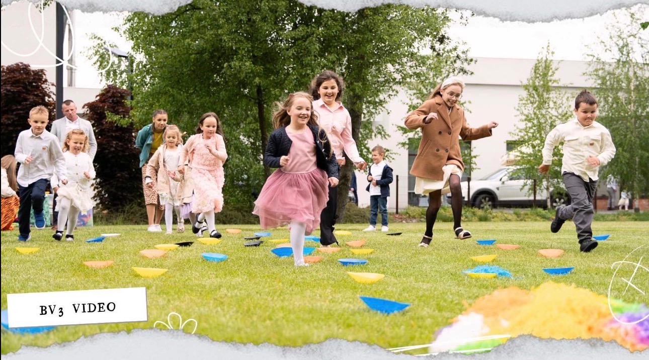 Grupa dzieci biegnie po trawie po kolorowych podkładkach. Widoczny kamerzysta nagrywający wydarzenie. Ujęcie z niskiego kąta, plenerowe, z zielenią w tle.
