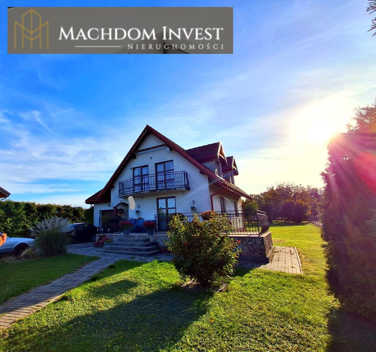 Dwupiętrowy dom z balkonem, z ogrodem i brukowanym chodnikiem, w słoneczny dzień. Logo firmy nieruchomości w górnej części kadru.