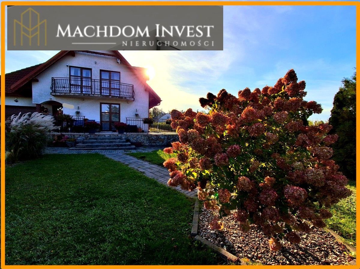 Dwupiętrowy dom z balkonem, widoczny z ogrodu z kwitnącymi hortensjami i kamienną ścieżką, pod błękitnym niebem. Logo firmy u góry kadru.