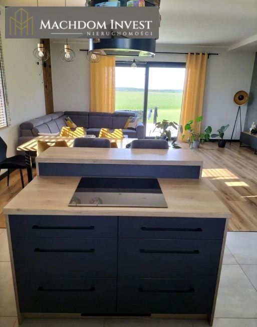 Nowoczesna kuchnia z wyspą i widokiem na zielone pola. Wnętrze domu z szarą sofą i żółtymi zasłonami. Jasne drewniane podłogi i minimalistyczne oświetlenie.