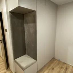 Szara szafa wnękowa z siedziskiem w przedpokoju, drewniana podłoga. Siedzisko z szarą tapicerką. Ściany w jasnym odcieniu szarości. Minimalistyczny design.