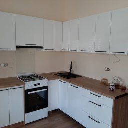 ProMebelek - Nowoczesna kuchnia z białymi, lakierowanymi frontami i drewnianym blatem. Widoczny zlew, kuchenka i wiszące szafki z czarnymi, podłużnymi uchwytami. Minimalistyczny design.